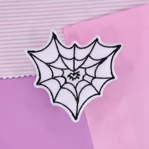 May include: A white felt spiderweb patch with black embroidered details. The spiderweb is in a heart shape. The patch is on a background of pink and lavender paper and fabric.