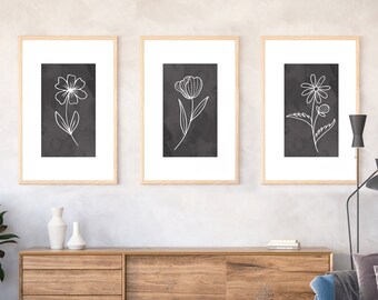 May include: Three framed black and white line drawings of flowers on a gray wall. The flowers are a daisy, a tulip, and a poppy. The frames are light wood.