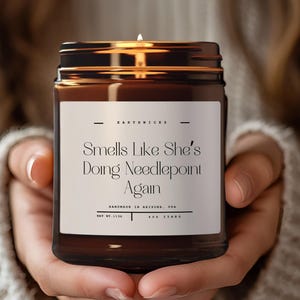May include: A lit candle in a brown glass jar with a black lid. The label reads "Smells Like She's Doing Needlepoint Again." The candle is held by a person wearing a cream-colored sweater. The candle is handmade in Arizona, USA.
