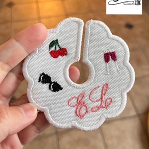 May include: White fabric wine glass charm with a scalloped edge, featuring embroidered cherries, a bow tie, and a wine glass. The charm has the embroidered initials "E Le" in pink and the text "Colours & Bobbins".