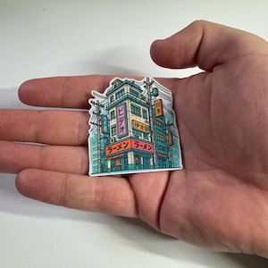 Puede incluir: Una colorida pegatina que representa un edificio de estilo japon&eacute;s con la palabra "Ramen" en japon&eacute;s. La pegatina tiene una combinaci&oacute;n de colores turquesa, rosa y naranja, con l&iacute;neas el&eacute;ctricas y otros edificios en el fondo.