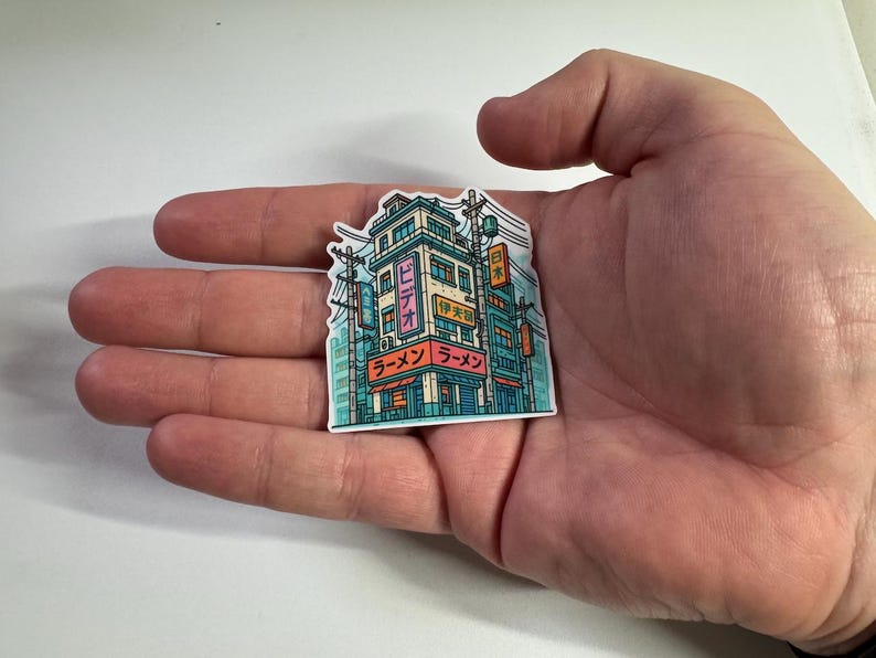 Puede incluir: Una colorida pegatina que representa un edificio de estilo japon&eacute;s con la palabra "Ramen" en japon&eacute;s. La pegatina tiene una combinaci&oacute;n de colores turquesa, rosa y naranja, con l&iacute;neas el&eacute;ctricas y otros edificios en el fondo.