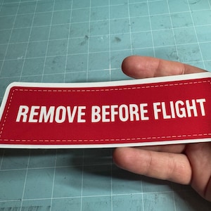 May include: A red rectangular sticker with the text "REMOVE BEFORE FLIGHT" in white, with a white border and a stitched design. The sticker is held in a hand.