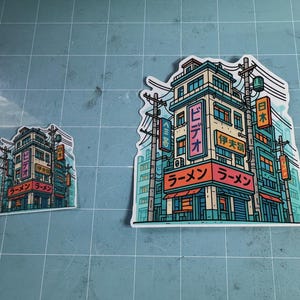 Puede incluir: Dos pegatinas con una colorida ilustraci&oacute;n de una tienda de ramen japonesa. El edificio es de varios pisos con letreros en japon&eacute;s. Las pegatinas son de estilo caricatura con acentos turquesa, naranja y rosa.
