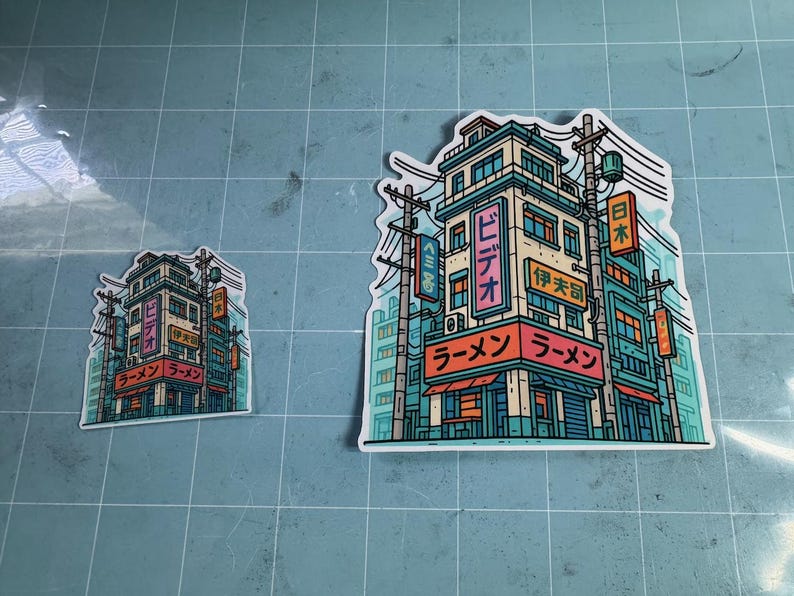 Puede incluir: Dos pegatinas con una colorida ilustraci&oacute;n de una tienda de ramen japonesa. El edificio es de varios pisos con letreros en japon&eacute;s. Las pegatinas son de estilo caricatura con acentos turquesa, naranja y rosa.