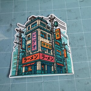 Puede incluir: Una colorida pegatina con una ilustraci&oacute;n estilizada de una tienda de ramen japonesa y los edificios circundantes. El dise&ntilde;o incluye texto japon&eacute;s y una vibrante paleta de colores de verde azulado, naranja y rosa.