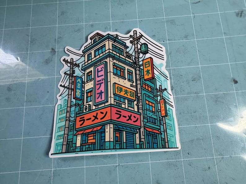 Puede incluir: Una colorida pegatina con una ilustraci&oacute;n estilizada de una tienda de ramen japonesa y los edificios circundantes. El dise&ntilde;o incluye texto japon&eacute;s y una vibrante paleta de colores de verde azulado, naranja y rosa.