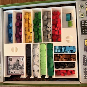 May include: A board game organiser with various game pieces in a green box. The pieces are sorted by colour, including blue, purple, yellow, green, grey, red, black, and brown. The game also includes cards with the number 38 on them.