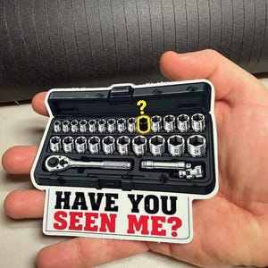 Puede incluir: Una pegatina rectangular que muestra un estuche de plástico negro para llaves de vaso con varias piezas de metal plateado, una carraca y una extensión. Un signo de interrogación amarillo resalta una pieza faltante, texto: HAVE YOU SEEN ME?