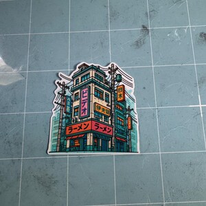 Puede incluir: Una pegatina con una ilustraci&oacute;n detallada de un edificio de estilo japon&eacute;s con la palabra "Ramen" en rojo y caracteres japoneses. El edificio es turquesa y beige con l&iacute;neas el&eacute;ctricas y letreros.