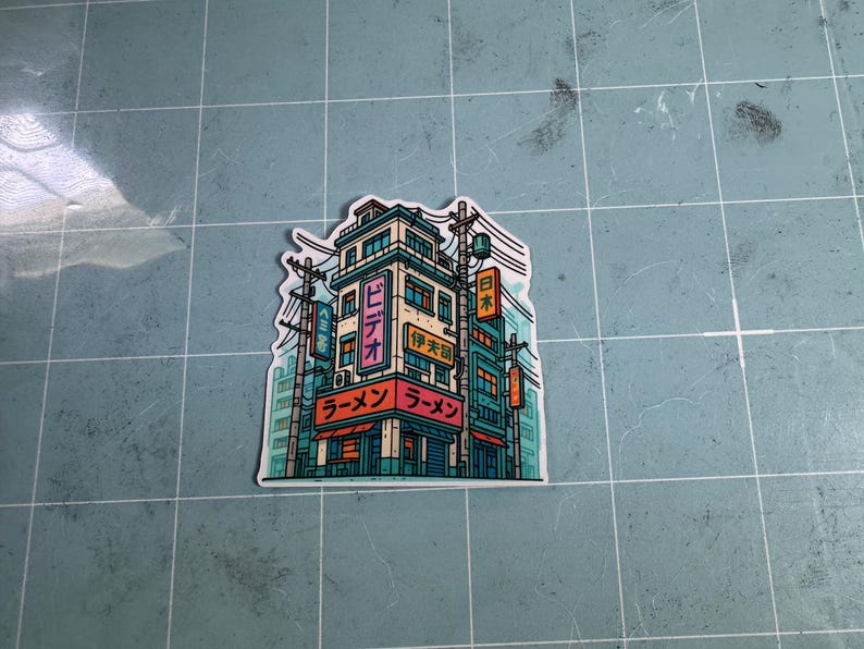 Puede incluir: Una pegatina con una ilustraci&oacute;n detallada de un edificio de estilo japon&eacute;s con la palabra "Ramen" en rojo y caracteres japoneses. El edificio es turquesa y beige con l&iacute;neas el&eacute;ctricas y letreros.