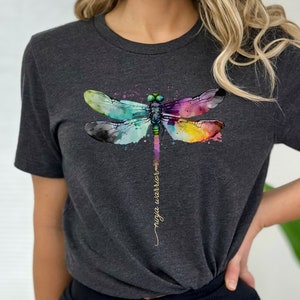 May include: A dark grey t-shirt with a watercolor dragonfly design in shades of blue, green, purple, yellow, and pink. The text "ninja warrior" is written vertically in gold script below the dragonfly.