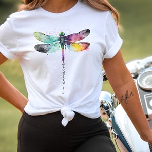 May include: White t-shirt with a watercolor dragonfly design and the word "warrior" written vertically down the center of the shirt.