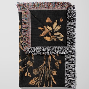 A woven throw blanket showcasing a majestic black dragon curled among mushrooms and botanical foliage beneath a glowing golden moon. The rich fantasy design, filled with forest plants and mystical details, is illustrated in earthy tones of crimson, gold, and deep brown on a dark backdrop.