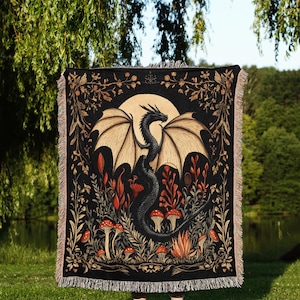A woven throw blanket showcasing a majestic black dragon curled among mushrooms and botanical foliage beneath a glowing golden moon. The rich fantasy design, filled with forest plants and mystical details, is illustrated in earthy tones of crimson, gold, and deep brown on a dark backdrop.