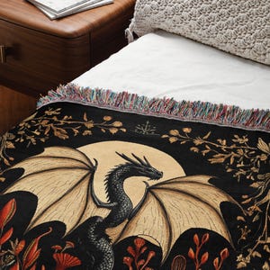 A woven throw blanket showcasing a majestic black dragon curled among mushrooms and botanical foliage beneath a glowing golden moon. The rich fantasy design, filled with forest plants and mystical details, is illustrated in earthy tones of crimson, gold, and deep brown on a dark backdrop.