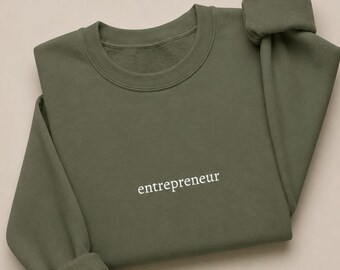 May include: An olive green crewneck sweatshirt featuring the word entrepreneur embroidered in white lowercase serif lettering across the center of the chest. The garment has long sleeves with ribbed cuffs and a ribbed neckline.