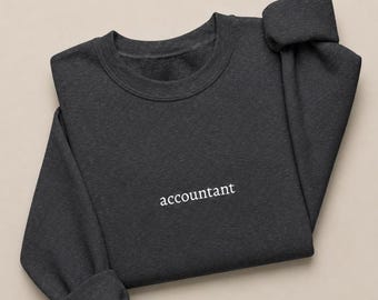 May include: A dark charcoal gray crewneck sweatshirt featuring the word accountant embroidered in white lowercase serif lettering across the center of the chest. The garment has long sleeves with ribbed cuffs and a ribbed neckline.