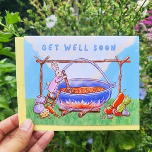 May include: A get well soon card featuring a cartoon bunny stirring a pot of soup over a campfire. The card has a blue sky background and the text "Get Well Soon" in a light blue font.