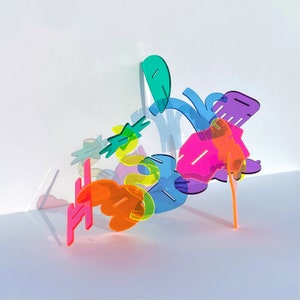 May include: Abstract sculpture composed of translucent, colorful acrylic shapes. The artwork features vibrant hues of pink, orange, yellow, green, blue, and purple. The forms are arranged in a dynamic, three-dimensional composition, casting shadows on a white surface.