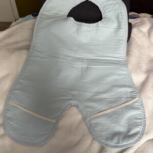 May include: A light blue baby bib with a curved neck opening and a wide, rounded bottom. The bib has a soft, quilted texture and features two white elastic bands near the bottom edges.