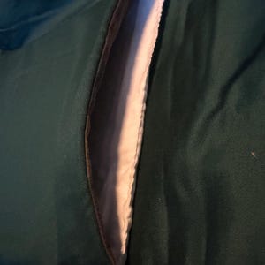 May include: Detail of a dark green jacket pocket. The pocket's interior is a light colour, with a brown trim. The jacket fabric has a subtle sheen, indicating a robust material, suitable for outerwear.