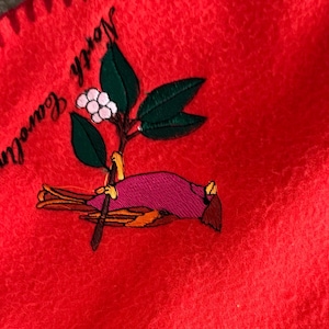 Puede incluir: Tela polar roja brillante con un diseño bordado. El diseño presenta un pájaro rosa, hojas verdes, flores blancas y las palabras "North Carolina" en una fuente curva.