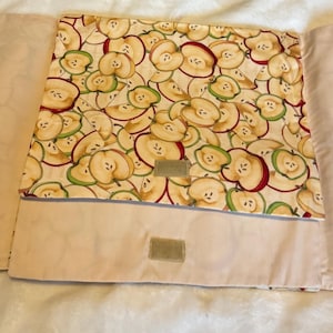 Puede incluir: Un artículo de tela beige con un estampado de rodajas de manzana. Las rodajas de manzana son rojas, verdes y amarillas. El artículo tiene un borde beige y un parche de velcro.