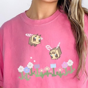 May include: Pink short-sleeved t-shirt with a graphic of two pixelated bees flying over a row of pixelated flowers. The bees are yellow and black, and the flowers are pink, purple, and blue.