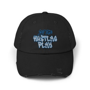Puede incluir: Gorra de béisbol negra desgastada con el texto "LET THE HUSTLAS PLAY" en azul claro y blanco. La gorra tiene una visera curva y un aspecto desgastado.