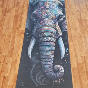 May include: A yoga mat with a colorful, detailed illustration of an elephant's face. The elephant's trunk is curled up and the background is black with white speckles. The text "THE PEACEMAKER" is at the bottom of the mat.