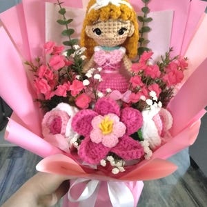 May include: A handmade bouquet featuring a crocheted doll with blonde hair and a pink dress, surrounded by pink and white flowers. The bouquet is wrapped in pink paper and tied with a white ribbon.