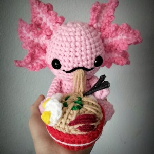 Peut inclure: Une figurine d'axolotl rose au crochet, avec des yeux noirs et une petite bouche. Il tient un bol de ramen avec divers garnitures. Le bol de ramen est rouge, beige et marron, avec des détails verts, blancs et rouges.