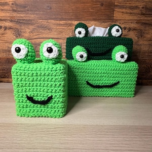 May include: Two handmade crochet tissue box covers in the shape of frogs. One is bright green, the other is dark green. Both have large, round eyes with white and black details, and a curved black smile. The tissue boxes are on a light brown surface.