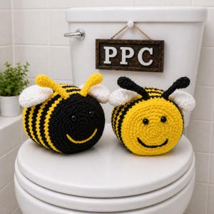 Puede incluir: Dos portarrollos de papel higiénico de abeja hechos a mano. Una abeja es negra y amarilla, la otra amarilla y negra. Ambas tienen alas blancas y caras sonrientes. Un letrero de madera con las letras "PPC" cuelga arriba.