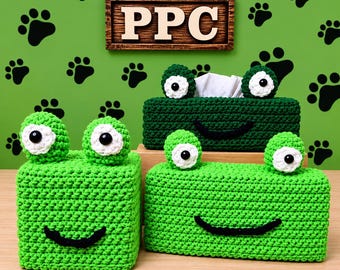 Könnte beinhalten: Drei grüne Häkel-Taschentuchbox-Abdeckungen in Froschform mit schwarzen gestickten Mündern sowie weiß-schwarzen Augen. Eine Abdeckung ist quadratisch, zwei sind rechteckig. Ein Holzschild mit der Aufschrift PPC hängt darüber.