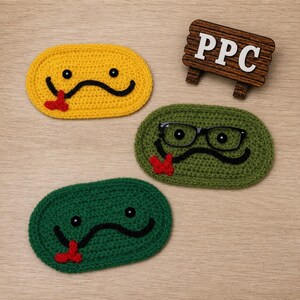 May include: Three handmade crochet coasters in yellow, green, and dark green, each with a smiling face design. The green coaster has black glasses. A small wooden sign with the letters "PPC" is also present.