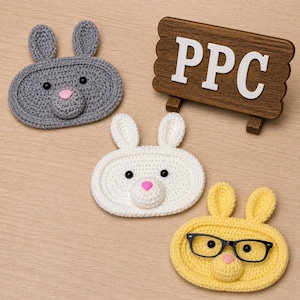 May include: Three oval-shaped crochet bunny faces in grey, white, and yellow, each featuring two ears, black bead eyes, and a pink nose. The yellow bunny wears black-rimmed glasses. A dark brown wooden sign with the white letters PPC is included.