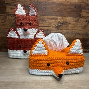 Puede incluir: Dos fundas de caja de pañuelos de ganchillo con forma de zorros. Una es naranja y blanca, con nariz y ojos negros, y la otra es marrón y blanca. Las fundas de la caja de pañuelos tienen orejas y narices de zorro.