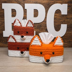 May include: Three rectangular crochet tissue box covers shaped like foxes. Two covers are dark orange and white with black eyes and noses, stacked vertically. One cover is bright orange and white with black eyes and a black nose, positioned in front.