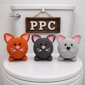 May include: Three crocheted cat-shaped toilet roll holders in orange, grey, and charcoal. Each cat has pink inner ears, a pink heart-shaped nose, and white whiskers. A wooden sign with the letters "PPC" hangs above.
