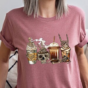May include: A pink t-shirt with a graphic of four coffee cups with skeletons holding them. The skeletons are wearing hats and have different poses. The text "Skeleton Crew" is printed on the shirt.
