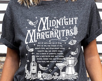 May include: A dark gray t-shirt with a white graphic design featuring a text overlay that reads "MIDNIGHT MARGARITAS" and includes a variety of illustrations, including a frog, a bat, a butterfly, a bottle of tequila, a cauldron, and a martini glass. The text below the graphic reads "Eye of newt, and toe of frog. Wool of bat, and tongue of dog. Adder's fork, and blind-worm's sting. Lizard's leg, and owlet's wing. For a charm of powerful trouble, Like a hell-broth boil and bubble. Double, double toil and trouble Fire burn, and caldron bubble."