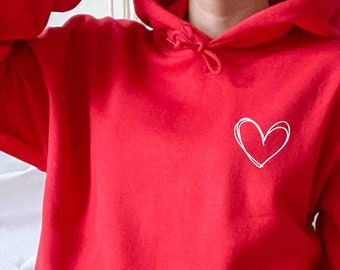 May include: A bright red hooded sweatshirt featuring a white hand-drawn heart outline on the left chest. The garment includes a front kangaroo pocket, a drawstring hood with matching red cords, and long sleeves with ribbed cuffs at the wrists.