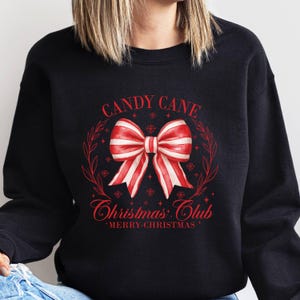 May include: Black sweatshirt with a red and white candy cane bow design. The text "CANDY CANE CHRISTMAS CLUB MERRY CHRISTMAS" is printed in red. The design is surrounded by a red wreath and snowflakes.