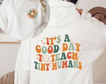 Może przedstawiać: Biała bluza z kapturem z małą grafiką kwiatowej uśmiechniętej buźki na lewej piersi oraz napisem "IT'S A GOOD DAY TO TEACH TINY HUMANS" dużymi, kolorowymi, falistymi literami na plecach, ozdobionymi małymi gwiazdkami.