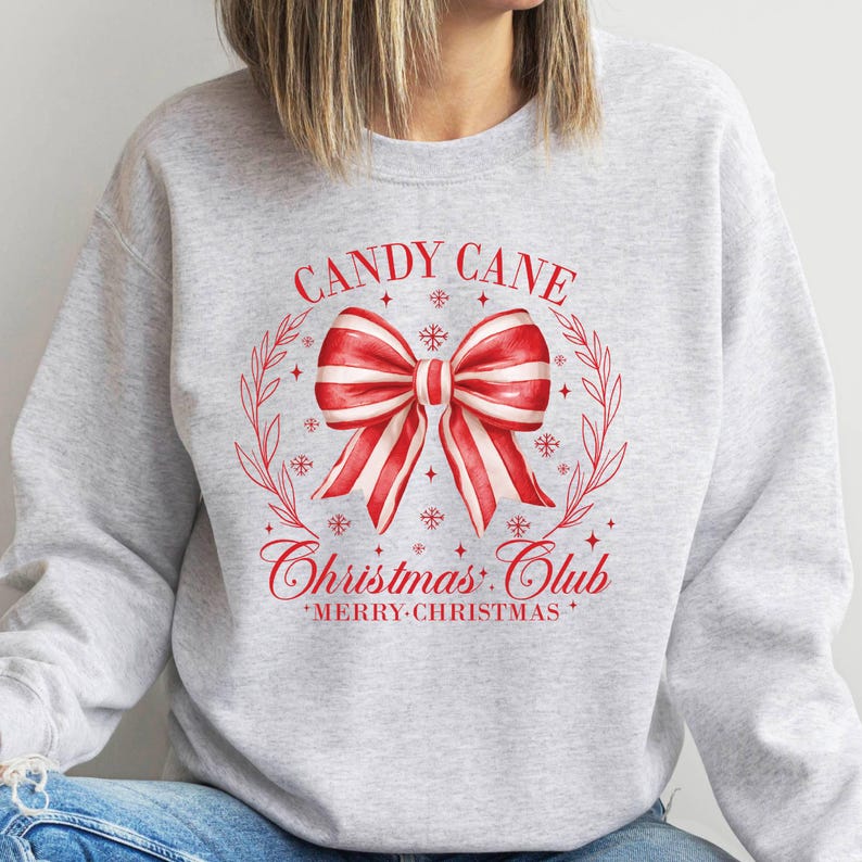 May include: A light grey sweatshirt with a red and white candy cane bow design. The text "CANDY CANE Christmas Club" and "MERRY CHRISTMAS" are printed in red. The design is surrounded by a wreath and snowflakes.