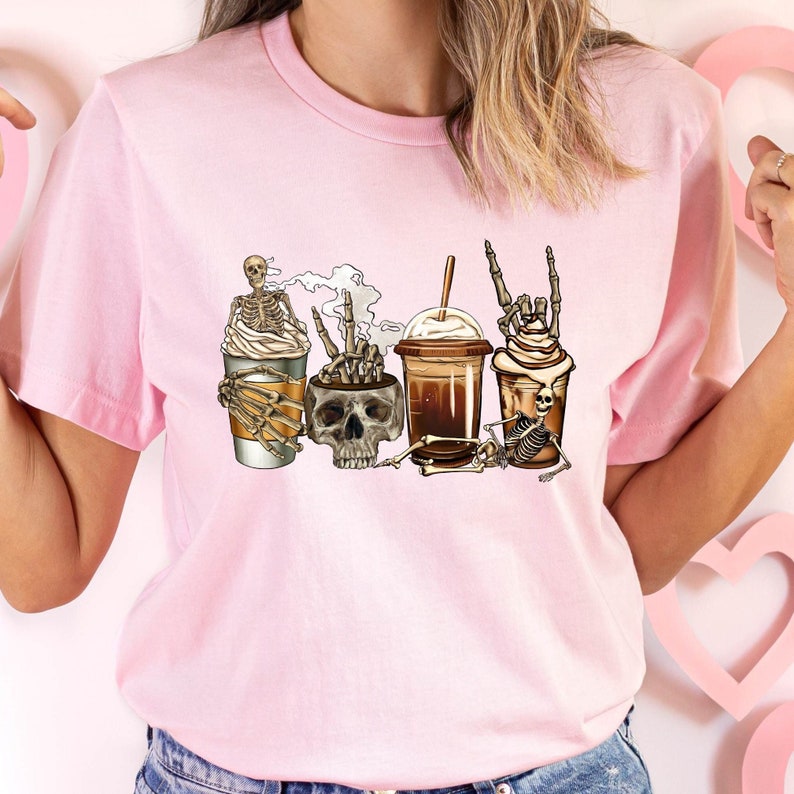 May include: Pink women's t-shirt featuring a whimsical design of skeletons enjoying various coffee drinks.  The illustration includes skeletons holding coffee cups, a skull in a coffee mug, and a skeleton with a frappuccino. The style is spooky and cute, perfect for Halloween or coffee lovers. The shirt is a light pink color and appears to be made of soft material.