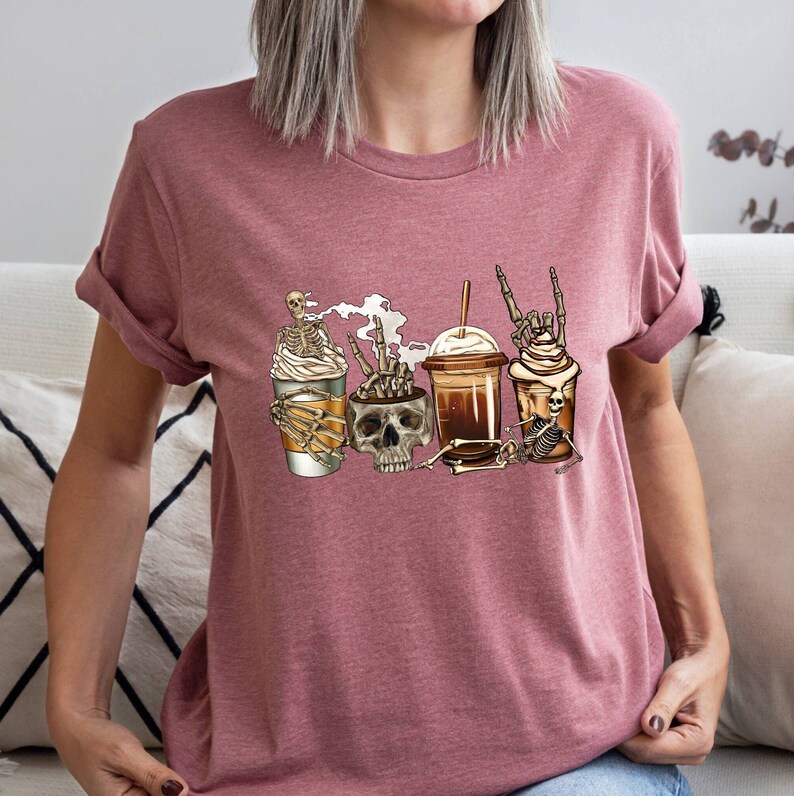 May include: A pink t-shirt with a graphic of four coffee cups with skeletons holding them. The skeletons are wearing hats and have different poses. The text "Skeleton Crew" is printed on the shirt.
