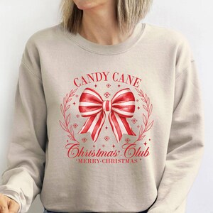 May include: A beige sweatshirt with a red and white candy cane bow design. The text "CANDY CANE Christmas Club" is printed above and below the bow, with "MERRY CHRISTMAS" at the bottom. The design is framed by a wreath.